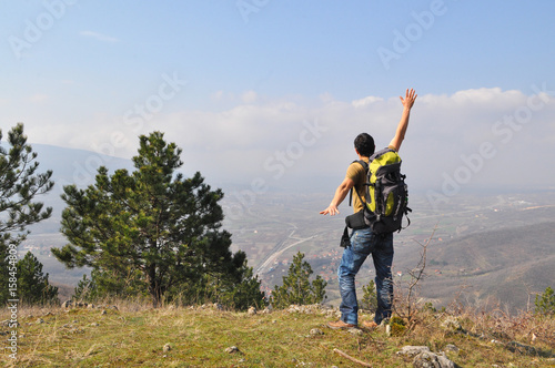 Young man with backpack standing on top of the hill over the city. Man spreads hands and enjoy in a wonderful view