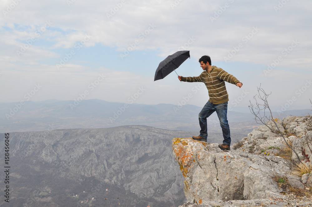 Obraz premium Young man with umbrella on a cliff edge on the top of mountain. Dangerous cliff on top of the mountain