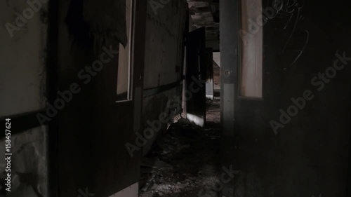 FPV, CLOSE UP: Walking along dark narrow hallway in abandoned decaying psychiatric hospital. Moving past the weathered wooden doors and broken crumbling walls in sinister sanatorium looking for escape photo