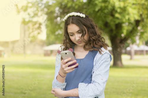 Beautiful and very young teenage girl standing in park and looking at smartphone