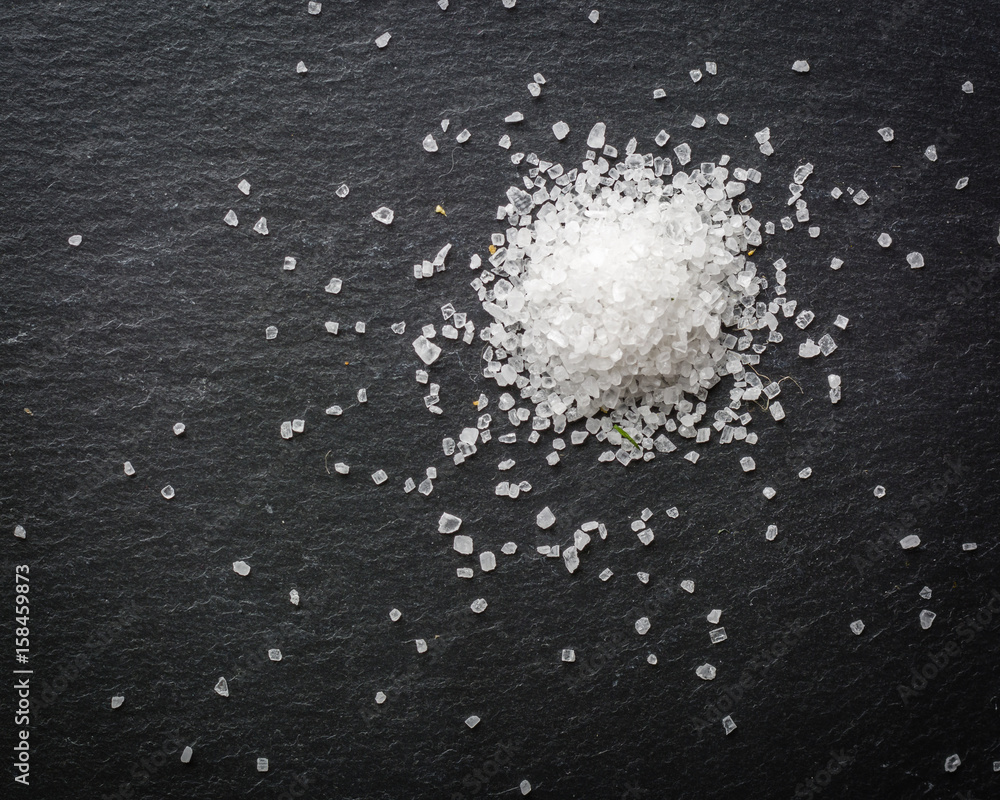 A bunch of white salt crystals on a black stone background. Seasoning for eating. Scattered over the surface. A bunch of white salt crystals on a black stone background. Seasoning for eating. Scattered over the surface.