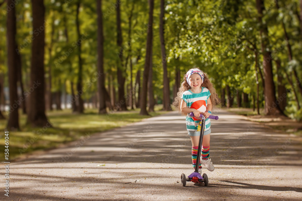 Fototapeta premium Little girl learn to ride scooter in a park on sunny summer day.