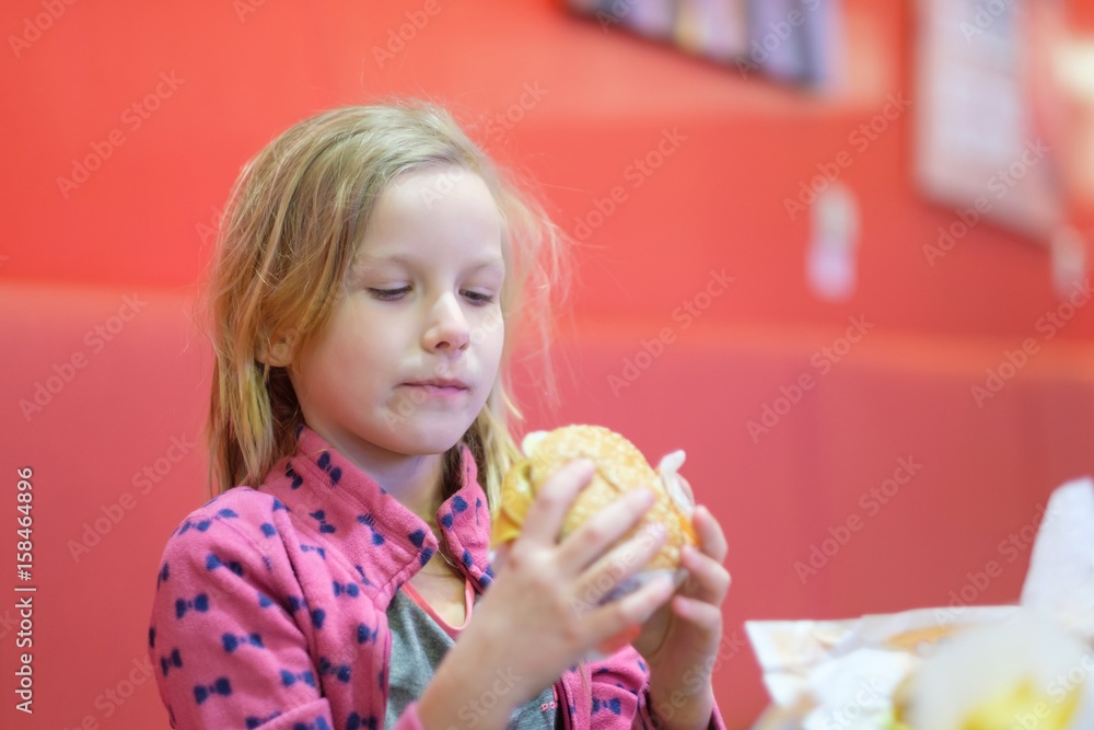 Adorable blonde girl in pink clothes eat double burger with beef and cheese in fast food restaurant Adorable blonde girl in pink clothes eat double burger with beef and cheese in fast food restaurant