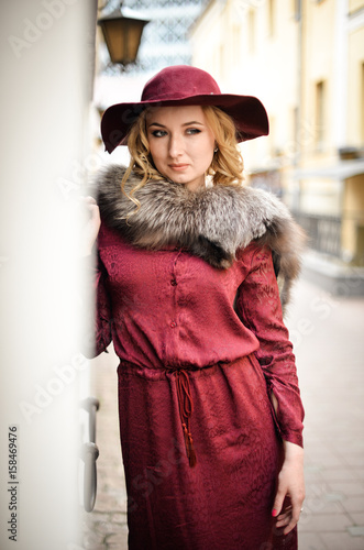 Красивая девушка со светлыми волосами стоит на улице в бордовом платье и шляпе с меховым воротником 
