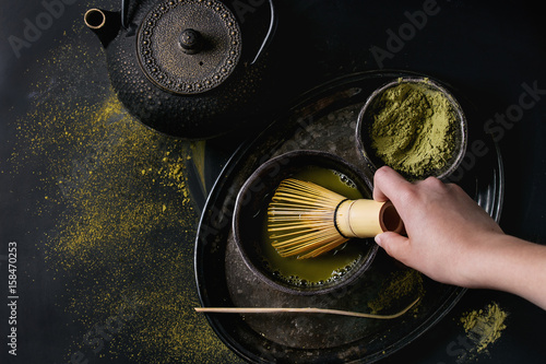Green tea matcha powder and hot drink in black bowls standing with iron teapot, bamboo traditional tools spoon and whisk in hand in vintage tray over dark metal background. Top view with space