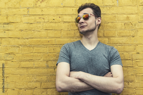 Pretty handsome confident young man hipster with sunglasses smiling behind yellow wall. Looking aside. Horizontal.