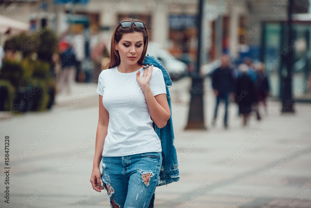 Attractive girl in a white T-shirt and jeans walking along the street. Mock-up.