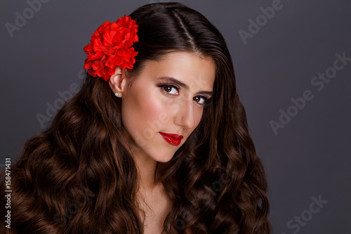 Beauty Fashion Model Woman face. Portrait with Red flower and red Lips. Beautiful Brunette Woman with Luxury Makeup and long hair.