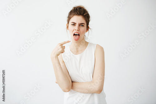 Photography Portrait of bored displeased young girl pointing finger at her tongue over white background