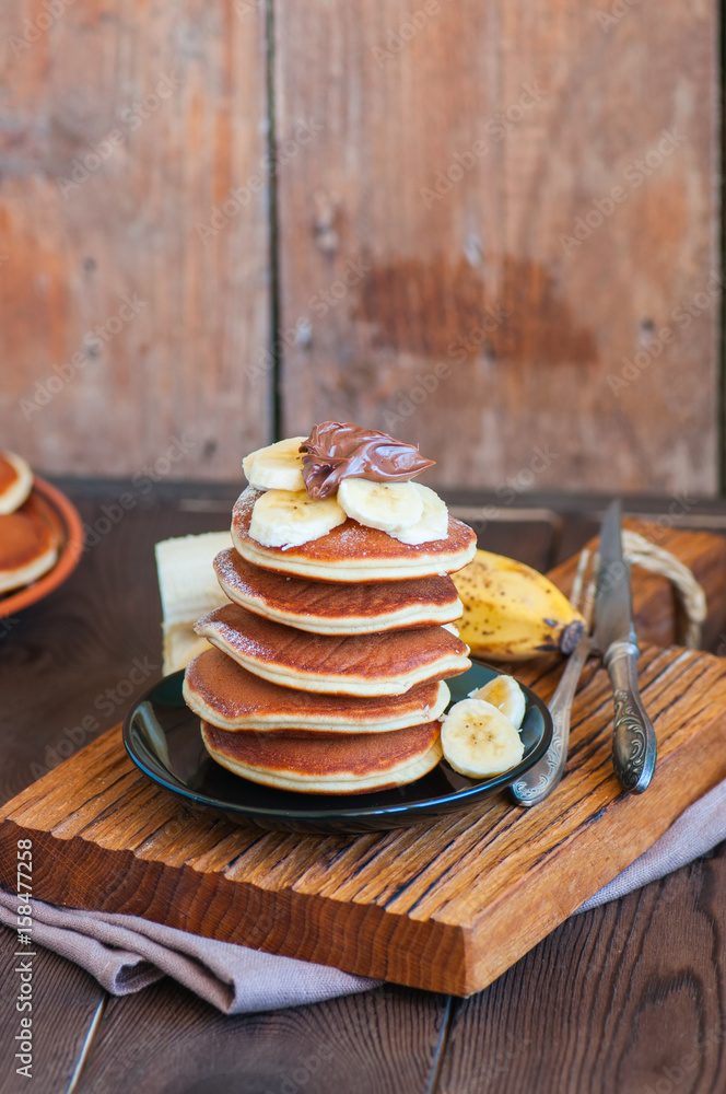 Close up of stack of homemade pancakes with bananas and chocolate paste in a plate on a board. Wooden background. Healthy breakfast and food concept. Close up of stack of homemade pancakes with bananas and chocolate paste in a plate on a board. Wooden background. Healthy breakfast and food concept.