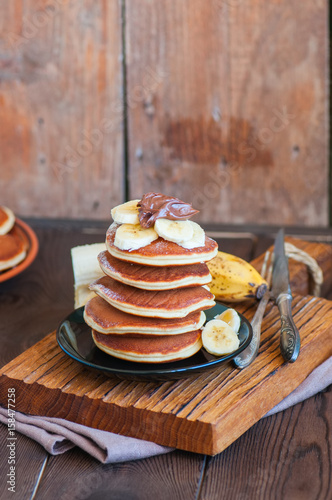 Close up of stack of homemade pancakes with bananas and chocolate paste in a plate on a board. Wooden background. Healthy breakfast and food concept.