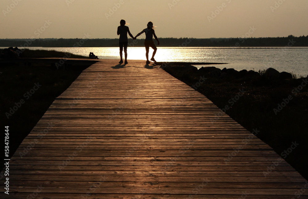 Fototapeta premium Two siblings walking along a boardwalk into the sunset.