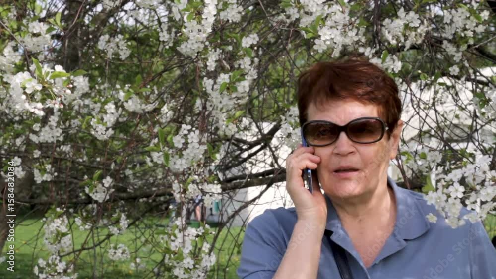 Medium shot of elderly woman wearing sunglasses standing in blossom, talking on mobile phone. Concept of modern technology used by senior adults Medium shot of elderly woman wearing sunglasses standing in blossom, talking on mobile phone. Concept of modern technology used by senior adults