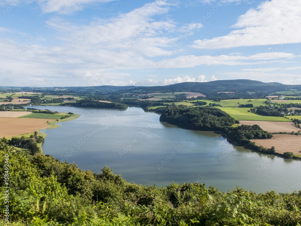 Panoramic view of Lake Huelgoat in Brittany, France. Around the lake you can see lush forests and cultivated fields. Blue sky with some clouds.