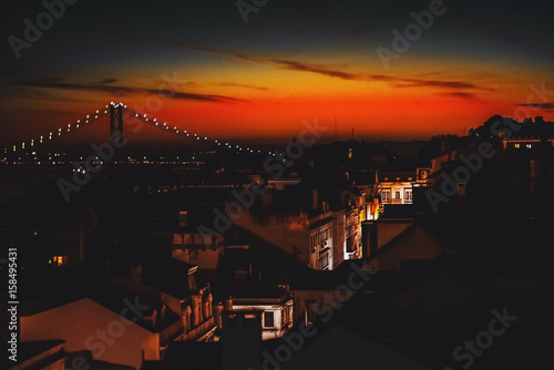 View from high point of illuminated narrow street with beautiful old facades of residential houses, following to rope bridge during stunning red and orange sunset in Lisbon, Portugal