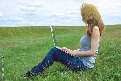 Woman sitting on a green meadow on the background of sky with clouds and working or studying with laptop wireless © Artem