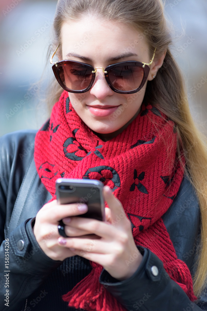 Fototapeta premium Beautiful female model wearing stylish leather coat and sunglasses is texting on her innovative smartphone. Urban blogger posting fashion and lifestyle news.