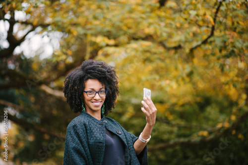 Happy young black woman taking selfie photo with smartphone on autumn. Stylish girl with afro hair using cellphone outdoor.