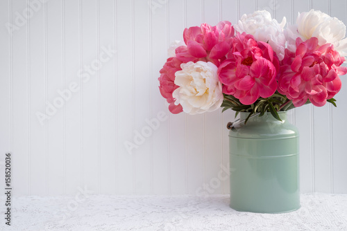 Pink and white peonies in a green vase on a white lace cloth in front of a white beadboard background.