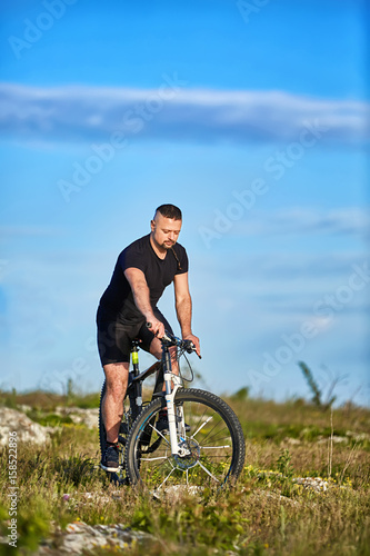 Attractive sportsman riding bike on rocky trail against sky with clouds.