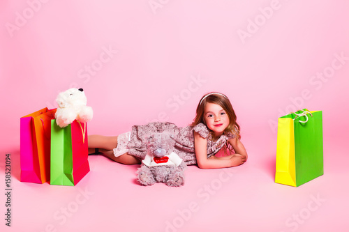 Little girl in a dress Lies on a pink background with a teddy bear and shopping bags, space for text photo
