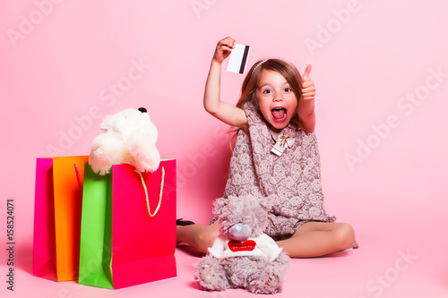 Little girl Shows thumb and tongue on a pink background with a teddy bear, shopping bags photo