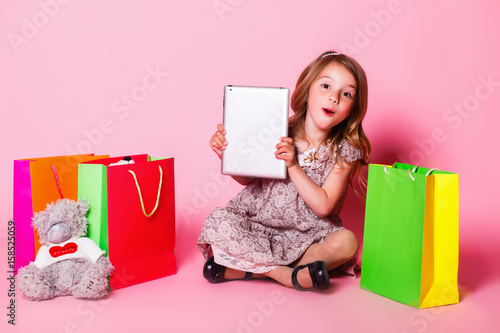 Little girl on a pink background with a teddy bear, shopping bags, and tablet fooling around
