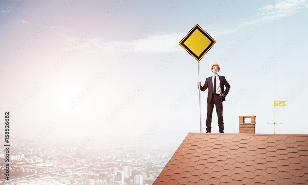 Young businessman on house brick roof holding yellow signboard and looking at city. Mixed media Young businessman on house brick roof holding yellow signboard and looking at city. Mixed media