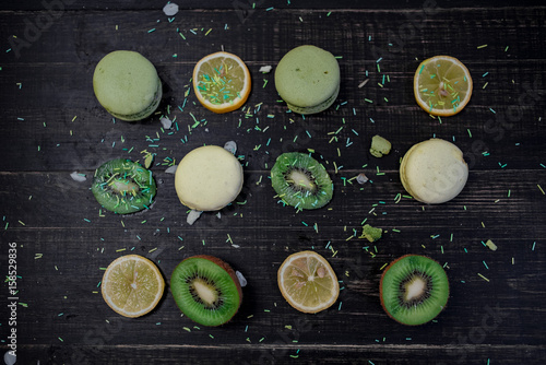Green and white macaroons with kiwi, orange and lemon in order on a wood gark table