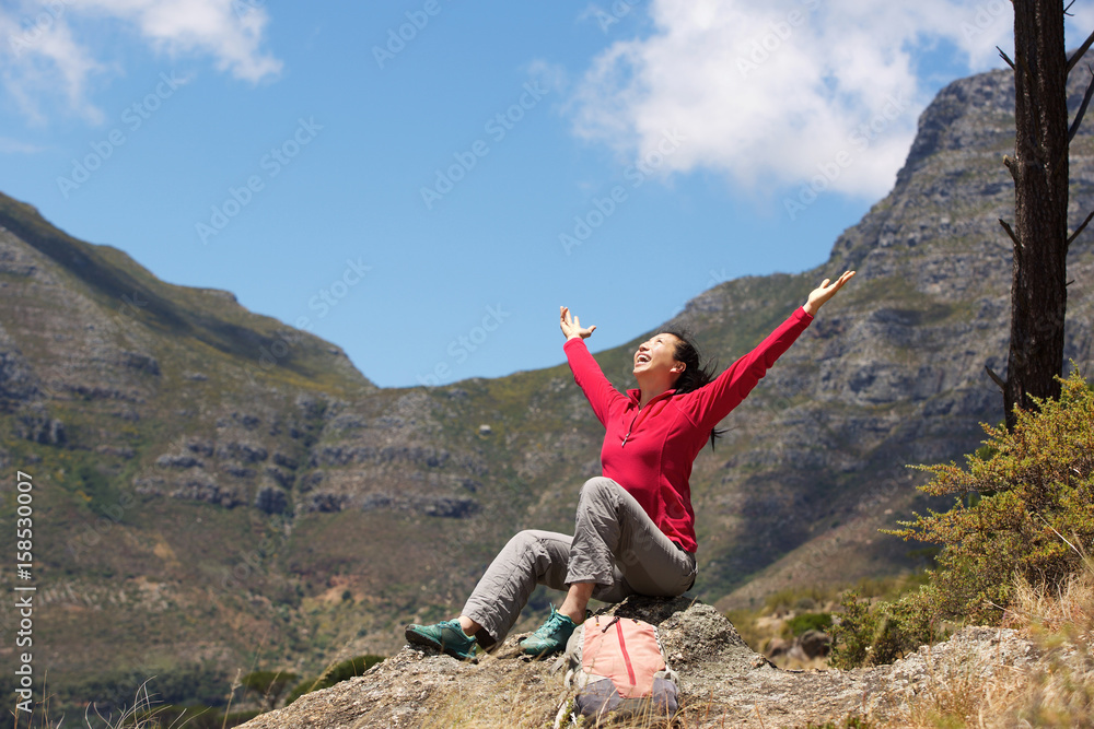 asian female hiker sitting on the peak with arms raised to the sky
