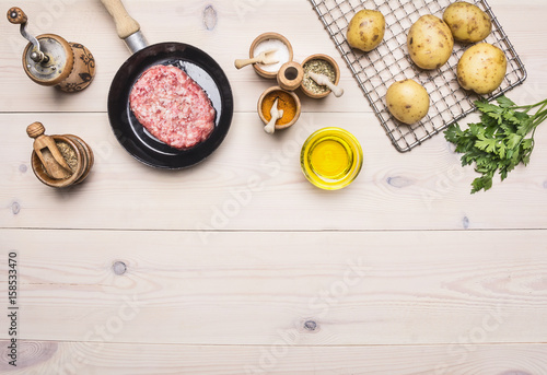 cooking homemade hamburger with fried potatoes, fresh ground beef in a small pan with parsley, different spices on white wooden table border place for text top view close up
