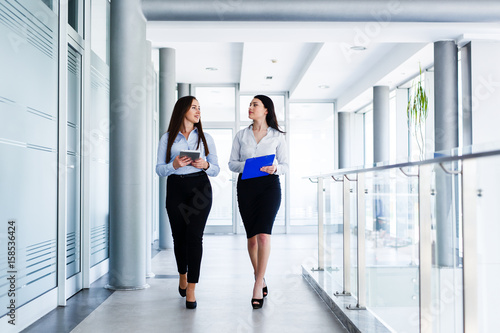Two attractive womens walking trought modern interior and talking about work