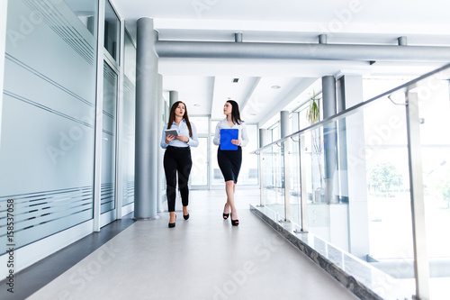 Two attractive womens walking trought modern interior and talking about work