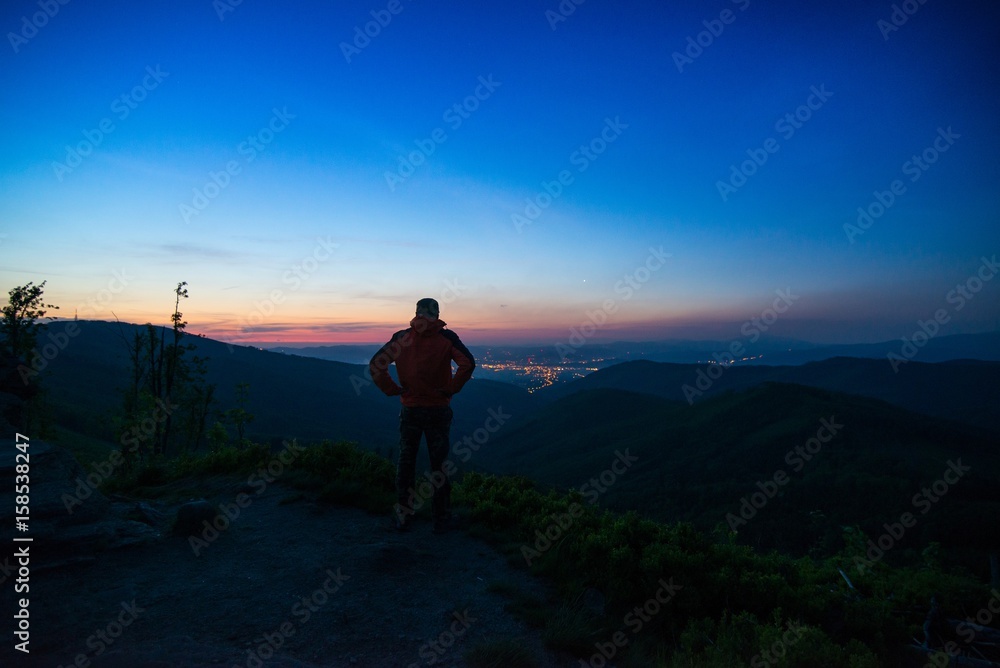 Man tourist standing on mountaintop observing city lights.