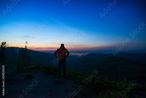 Man tourist standing on mountaintop observing city lights.