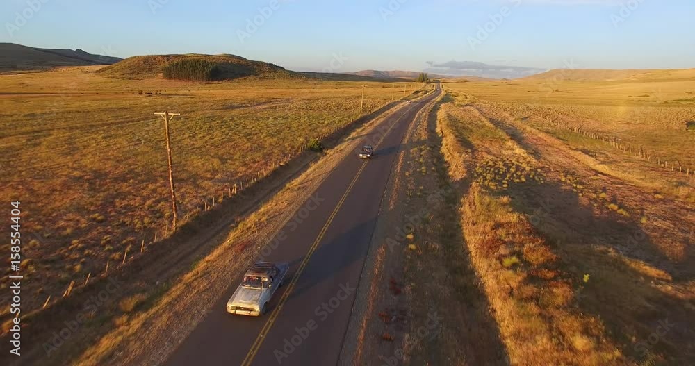 Aerial drone scene of sunset golden hour in Patagonia, Argentina. Steppe landscape. Camera moving backwards fast tracking two cars on the road. One is a old vintage car. Aerial drone scene of sunset golden hour in Patagonia, Argentina. Steppe landscape. Camera moving backwards fast tracking two cars on the road. One is a old vintage car.