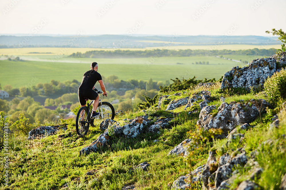 Rear view of the young cyclist riding the bike on the rocky trail in summer.