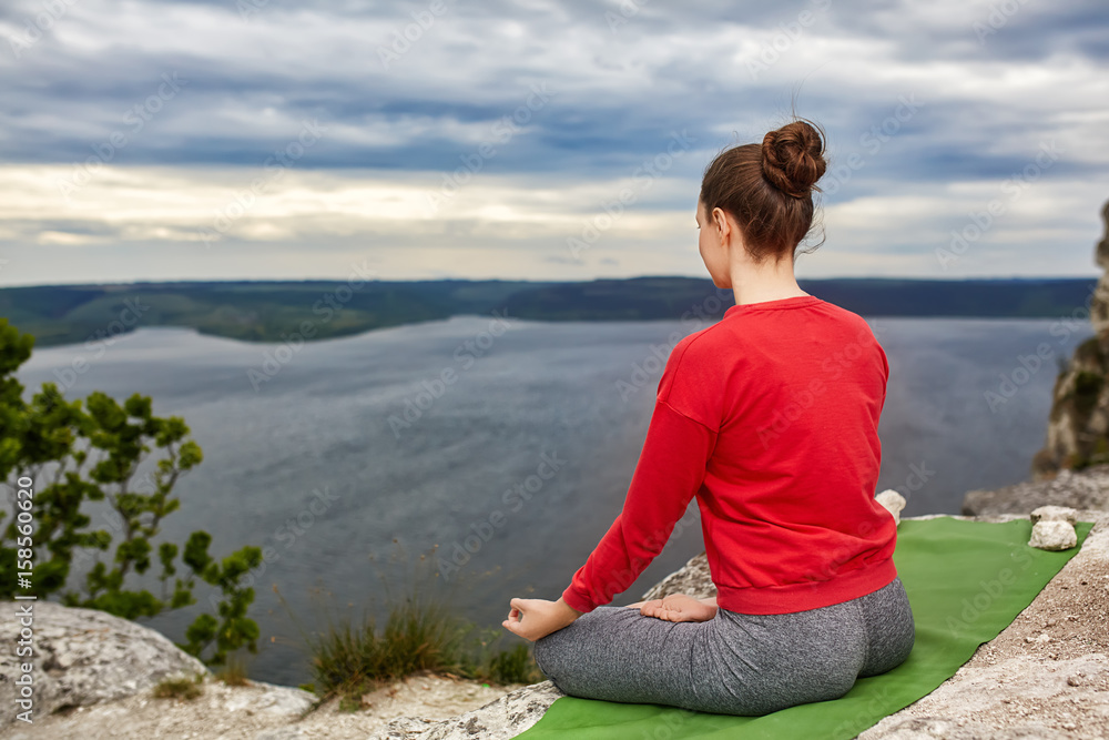Young woman meditating in a lotus pose on the rock above big river.