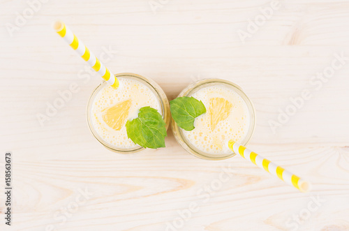Freshly blended yellow lemon smoothie in glass jars with straw, mint leaf, top view. White wooden board background, copy space.