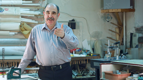 Portrait of a senior male business owner behind the counter of his frame workshop, standing and talking to a camera