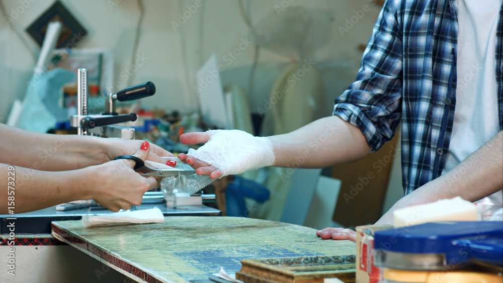 Young female colleague carefully bandaging worker injured hand after accident in workshop Young female colleague carefully bandaging worker injured hand after accident in workshop