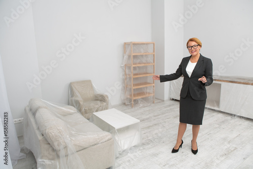 Woman realtor proposes to visit flat or apartment. Agent in glasses shows the living room of an apartment.