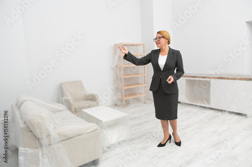 Realtor in living room proposes to view a flat - real estate. Mid aged woman standing in the centre of lthe room, showing the apartment. photo