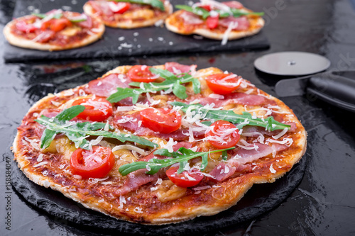 Homemade pizza with prosciutto, tomato, arugula on black slate board. Close up.