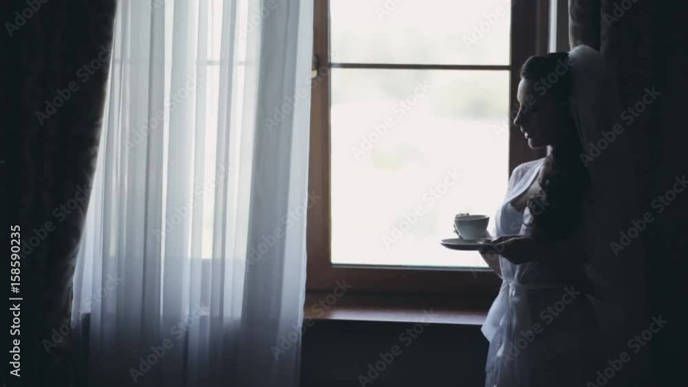 Young bride staying near the window and drinking coffee in morning. Woman with veil getting ready to wedding ceremony. Young bride staying near the window and drinking coffee in morning. Woman with veil getting ready to wedding ceremony.