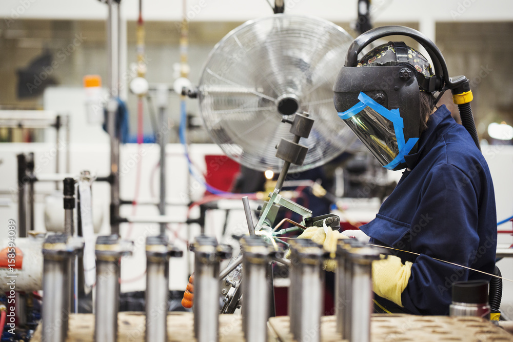 A person wearing a protective helmet using a welding torch in the assembly process in a cycle factory. A large fan cooling the heated metals. A person wearing a protective helmet using a welding torch in the assembly process in a cycle factory. A large fan cooling the heated metals.