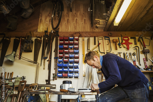 Man standing in a garden workshop surrounded by tools hanging from beams and a stack of garden tools propped up.
