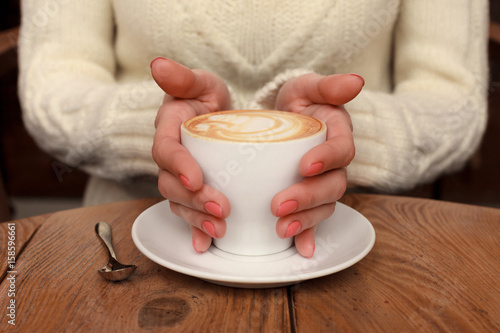 Close-up female hands holding cup with coffee cappuccino with foam with nice pattern. Perfect red gel polish manicure. Wood natural vintage table. Creative color warm post processing