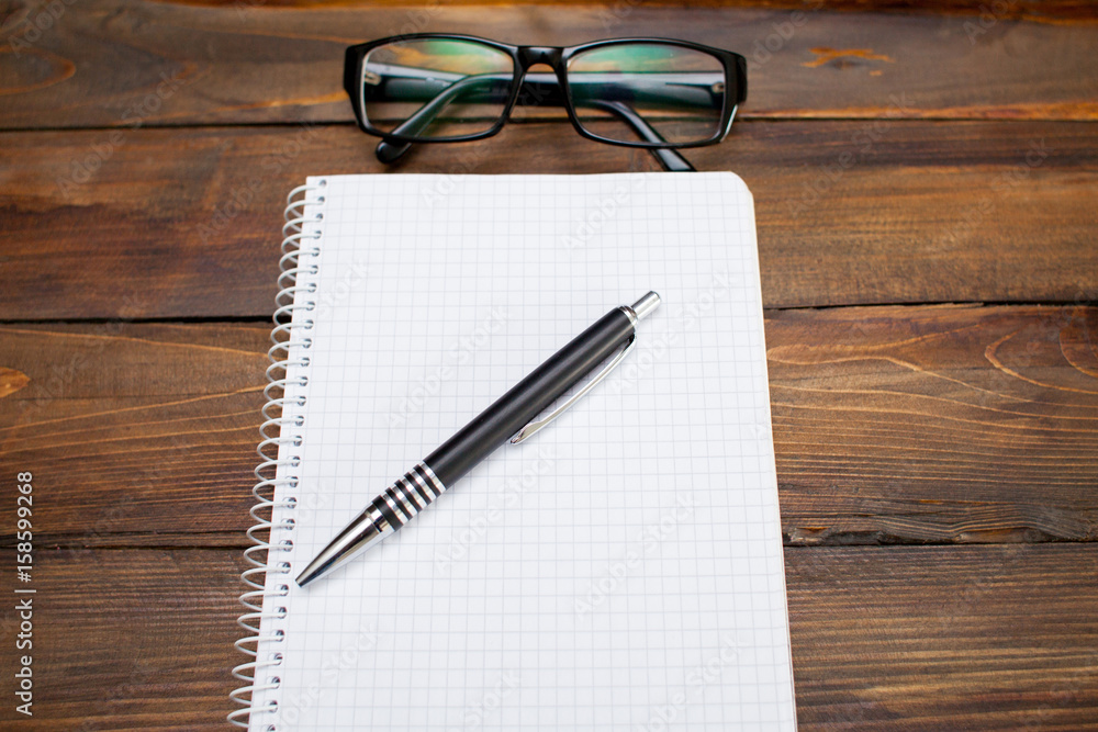 Fototapeta premium Notepad in box with white pages, glass spectacles in black frame and pen on wooden background. Top view