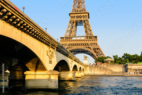 The Eiffel tower seen from the Debilly pier in a warm light at sunset with the Iena bridge and the river Seine in the foreground.